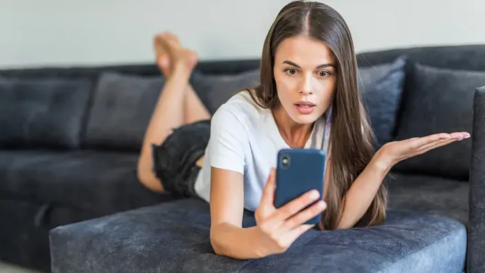 Joven confundida mirando su smartphone en el sofá debido a una mala cobertura o conexión a internet.