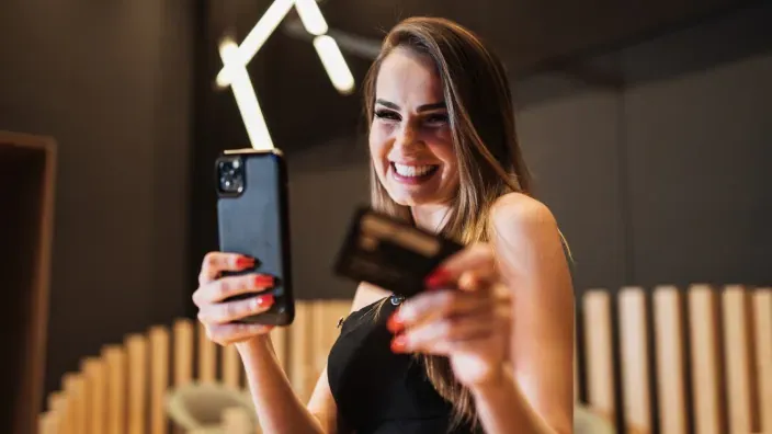 Mujer sonriendo mientras sostiene un móvil y una tarjeta bancaria