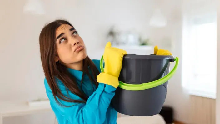 Mujer frustrada con un cubo por una gotera en el techo, representando siniestros protegidos por el seguro de hogar.
