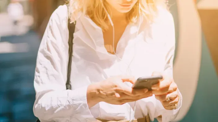 Mujer usando el móvil por la calle con auriculares durante una llamada o consulta online.