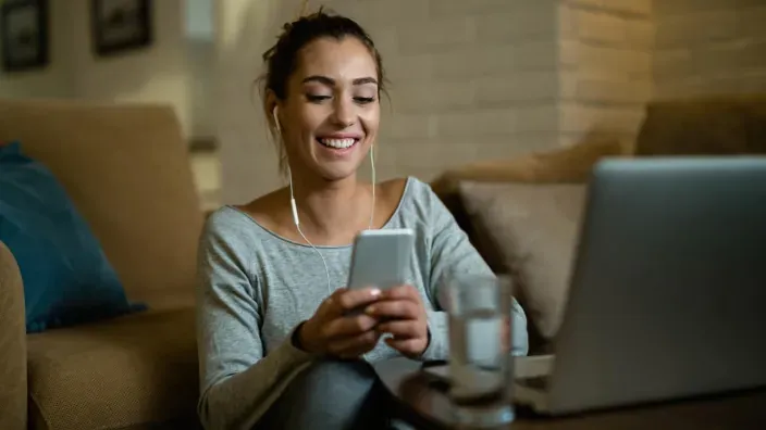 Joven sonriente con auriculares usando su smartphone junto a un portátil en el salón de casa.
