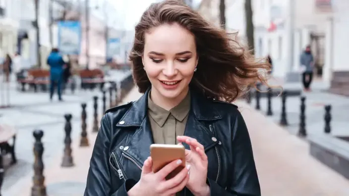 Mujer sonriente usando su smartphone mientras camina por una calle peatonal para representar la conectividad móvil.