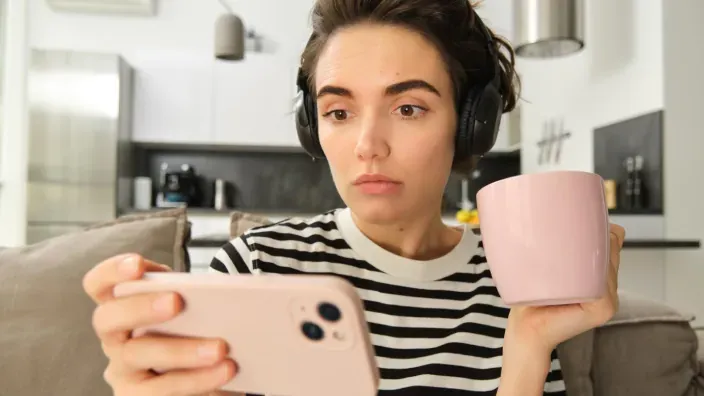 Mujer con auriculares y taza en la mano mirando su móvil con expresión de sorpresa.