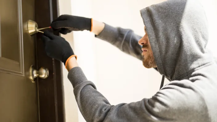 Ladrón con sudadera y guantes forzando la cerradura de una puerta con una ganzúa para entrar a robar a una casa.