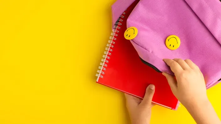 Niño guardando un cuaderno rojo en una mochila rosa sobre un fondo amarillo, preparándose para la vuelta al cole.