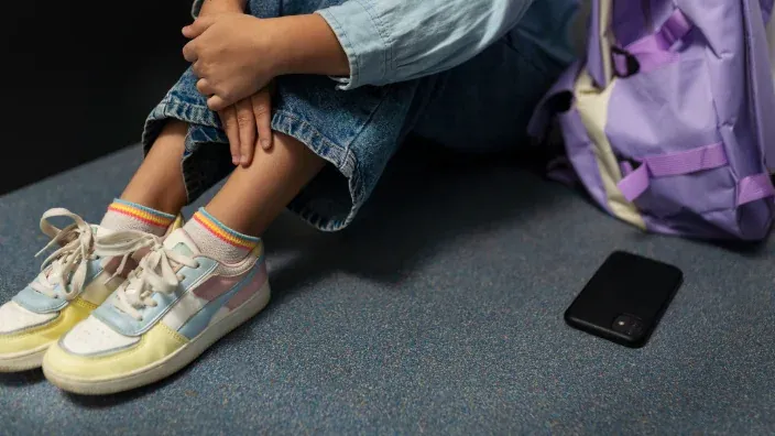 Estudiante sola y triste en el suelo del colegio junto a su móvil. Símbolo del ciberacoso (bullying) y la salud mental infantil.