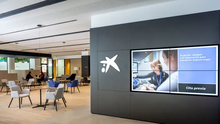 Interior de oficina Store de CaixaBank con pantalla digital de cita previa y mobiliario moderno.