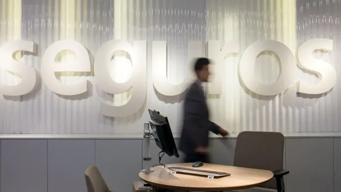 Oficina de seguros con un gran logotipo corporativo en la pared y una persona pasando delante de un escritorio