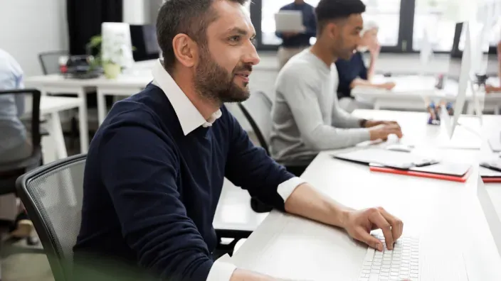 Empleado trabajando frente al ordenador en una oficina moderna con compañeros al fondo.