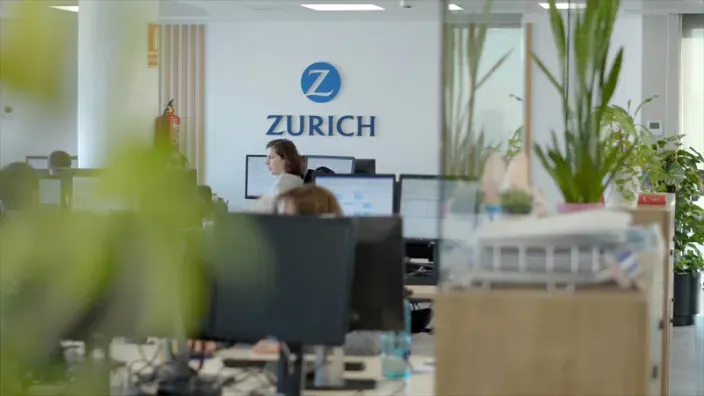 Interior de oficina de Zurich con empleados trabajando frente a ordenadores y logotipo de la compañía al fondo.