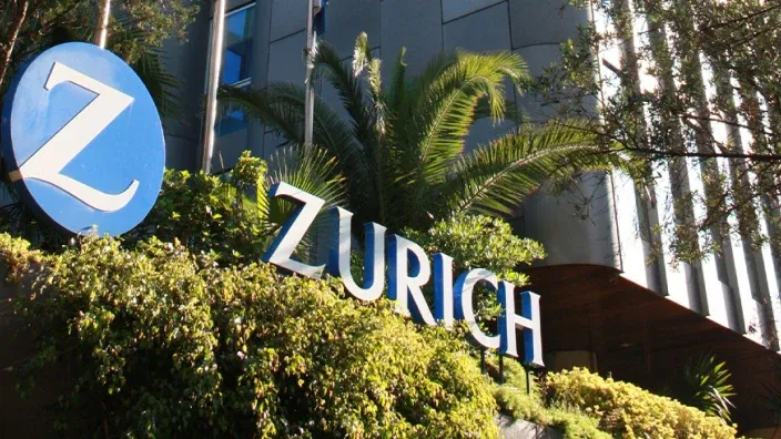 Logotipo de Zurich Seguros en un edificio de oficinas, rodeado de vegetación, con el icónico distintivo azul con la Z.