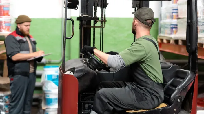 Mozo de almacén conduciendo carretilla elevadora mientras supervisor revisa inventario al fondo.