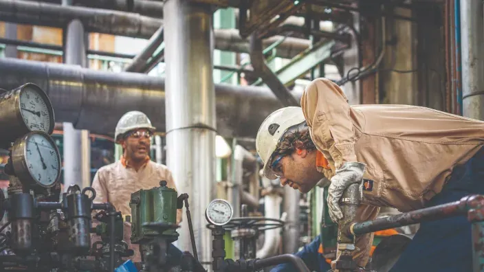 Dos operarios con casco trabajando entre tuberías y manómetros en una planta industrial