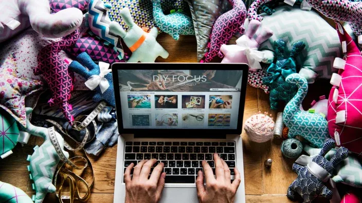 Ordenador portátil rodeado de telas, muñecos de tela y materiales de costura