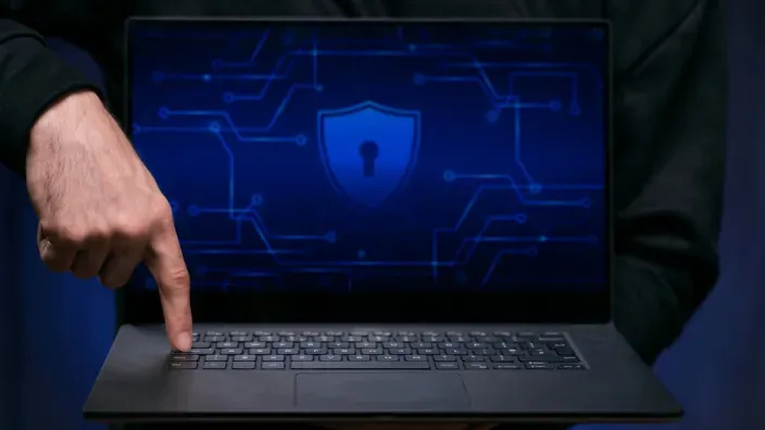 Mano de técnico pulsando tecla enter en portátil con escudo de seguridad azul en pantalla.