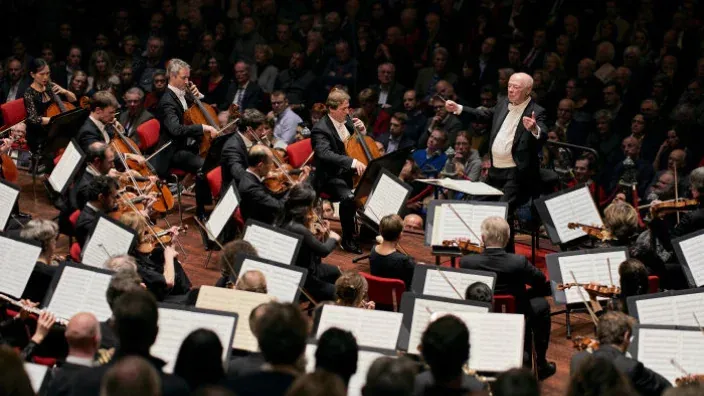 Orquesta sinfónica interpretando concierto de música clásica con director y violonchelistas.