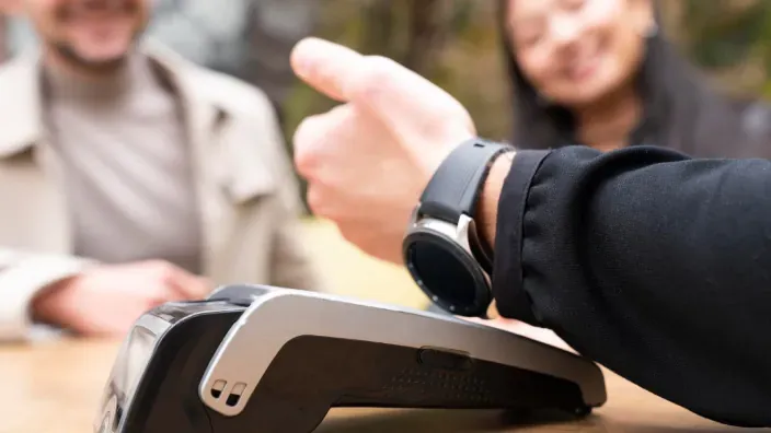 Persona acercando un reloj inteligente a un terminal de pago mientras dos personas observan al fondo