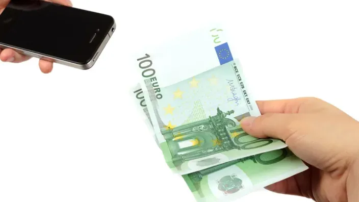 Mano entregando billetes de 100 euros junto a un teléfono móvil, representación de pago o transferencia de dinero.