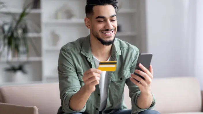 Joven usando tarjeta de crédito y smartphone para compras online o gestiones de banca digital desde casa.