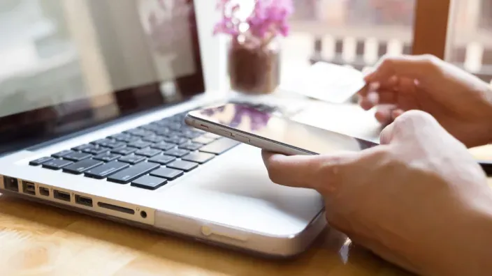 Persona realizando un pago online con el móvil y un portátil sobre una mesa de madera