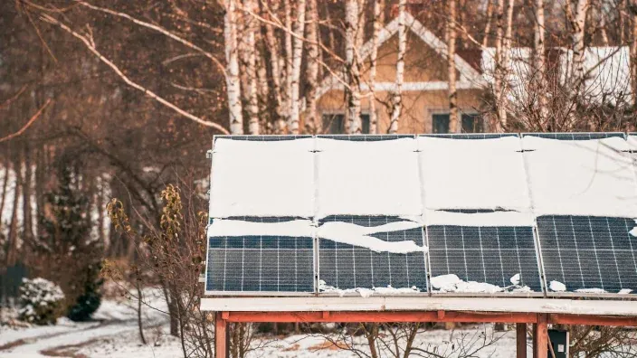 Paneles solares cubiertos parcialmente de nieve en invierno frente a una casa de madera.