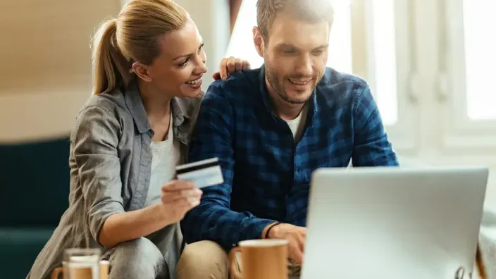 Pareja sonriente realizando compras online con tarjeta de crédito y portátil en casa para gestionar sus finanzas.