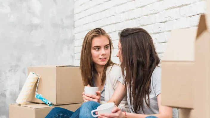 Dos mujeres jóvenes sentadas entre cajas de mudanza conversando y tomando café.