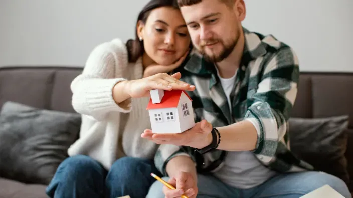Pareja joven protegiendo maqueta de casa con las manos simbolizando hipoteca.