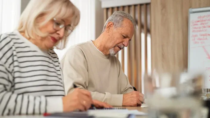 Pareja de jubilados escribiendo y revisando papeles financieros en una mesa durante la planificación económica.