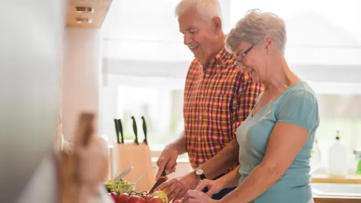 Pareja de jubilados sonriendo mientras cocinan juntos. 