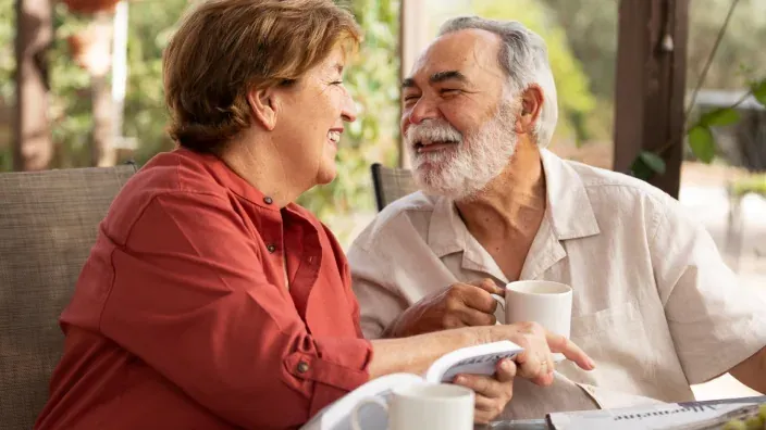 Pareja mayor sonriendo mientras toma café y revisa un libro en una terraza, imagen relacionada con la jubilación y el bienestar económico.