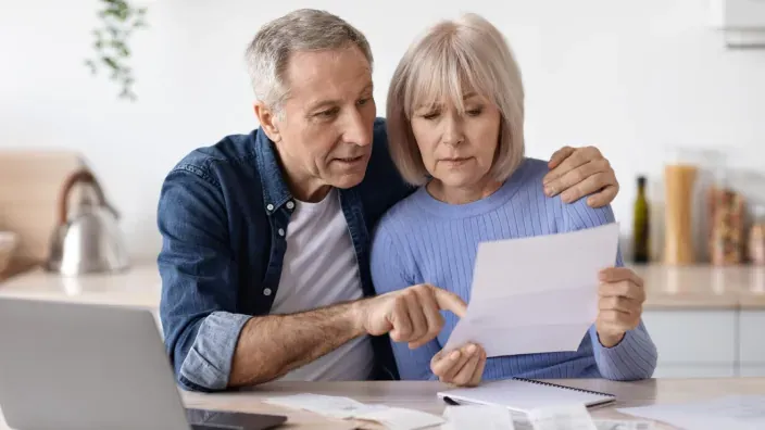 Pareja de jubilados revisando una carta y facturas en casa con un portátil para gestionar sus ahorros y pensiones.
