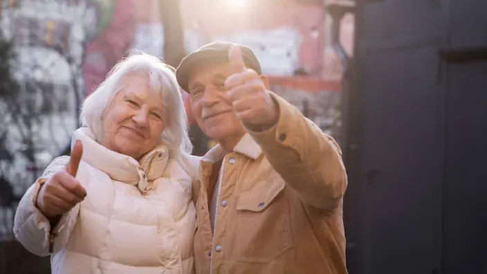 Pareja de personas mayores sonriendo y haciendo gesto de aprobación con el pulgar hacia arriba en un entorno exterior.