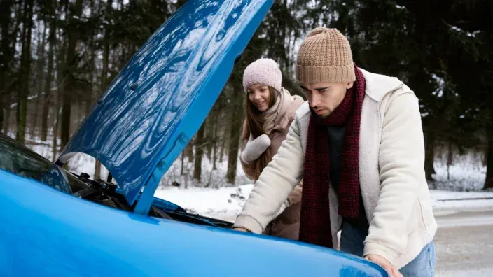 Joven revisando el motor de un coche en una carretera nevada mientras su acompañante observa.