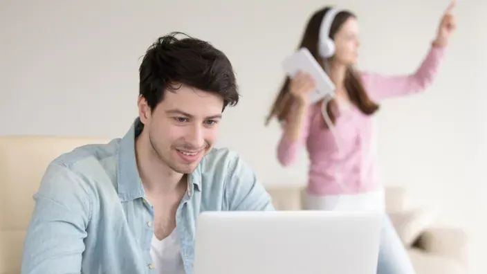 Hombre teletrabaja en su portátil mientras su pareja baila detrás con una tablet y cascos.