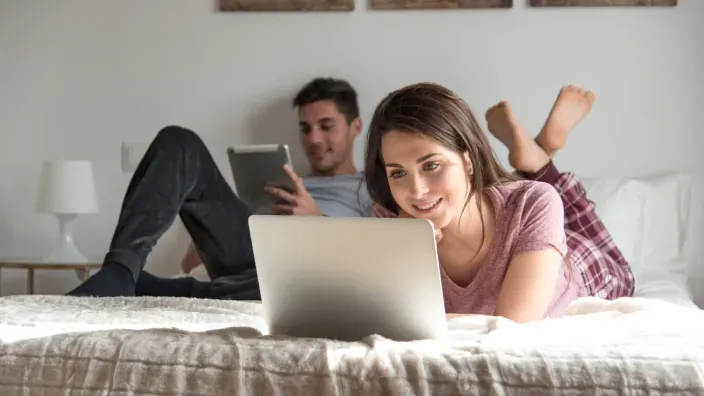 Pareja joven utilizando un portátil y una tablet en la cama para navegar por Internet o ver contenidos online.