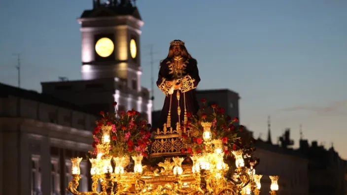 Paso de Semana Santa con una imagen de Cristo iluminada de noche y la torre del reloj de Sol en Madrid al fondo.