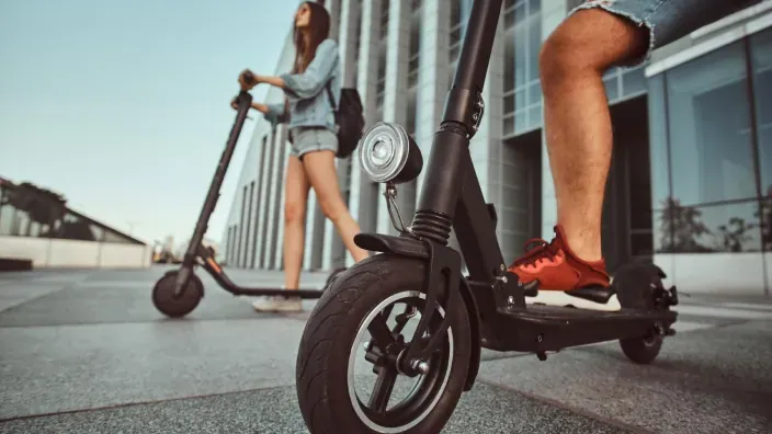 Dos personas utilizando patinetes eléctricos frente a edificios modernos en una zona urbana.