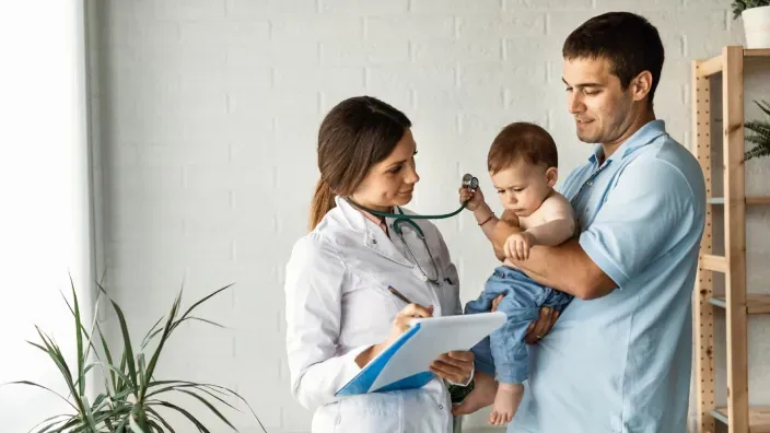 Doctora pediatra examinando a un bebé en brazos de su padre durante una revisión médica de un seguro de salud.