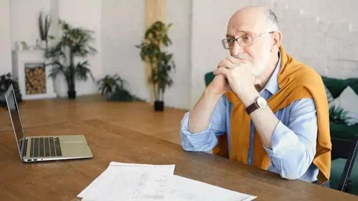 Hombre mayor revisando documentos financieros frente a su ordenador en casa, preocupado por su pensión.