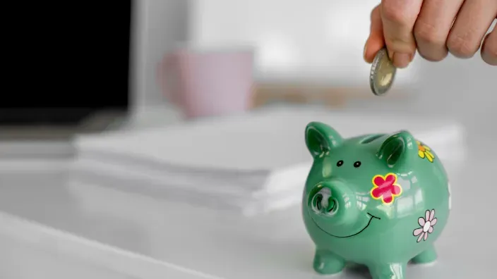 Una mano introduce una moneda en una hucha de cerdito verde, simbolizando el ahorro, la inversión y la gestión de finanzas.