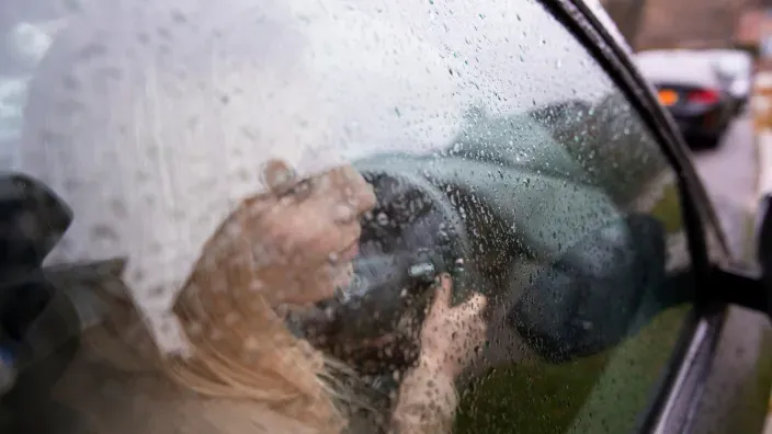 Conductora estresada o cansada, apoyada en el volante de su coche en un día de lluvia.