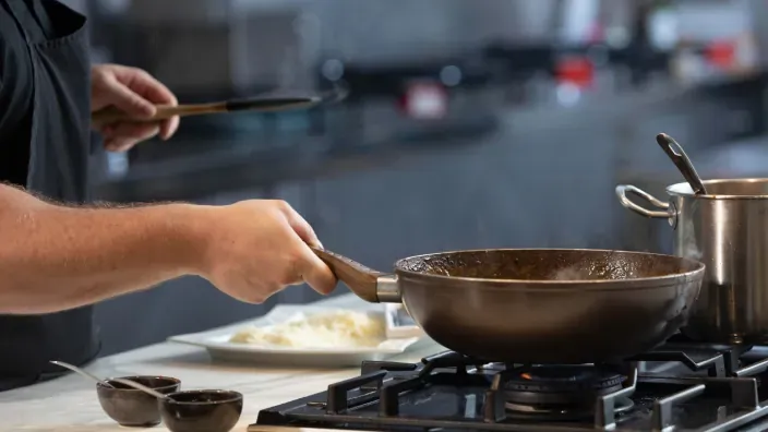 Cocinero utilizando una sartén sobre una cocina de gas en funcionamiento, preparando alimentos en casa.