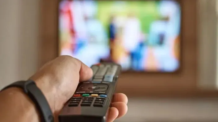 Primer plano de una mano sosteniendo un mando a distancia frente a un televisor encendido.