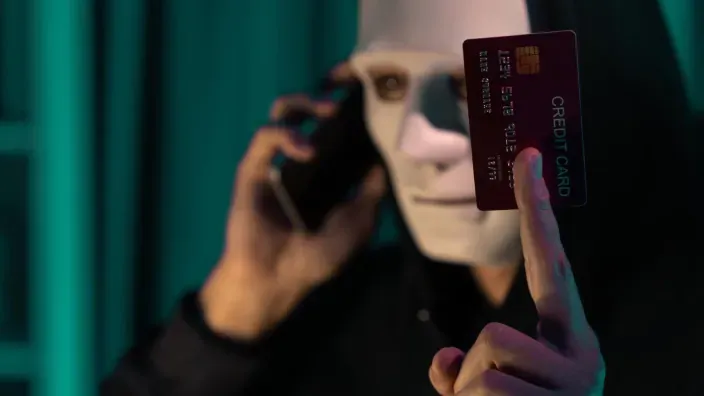 Persona con máscara blanca hablando por teléfono y sosteniendo una tarjeta bancaria