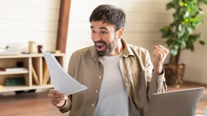 Hombre celebrando ahorro en factura con documento en la mano frente al portátil.