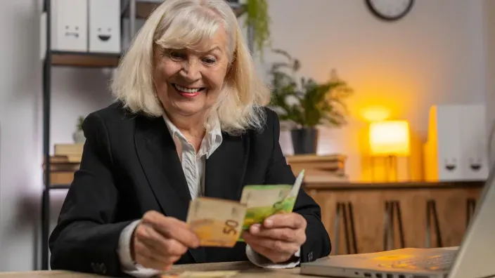 Mujer mayor sonriente contando billetes de 50 y 100 euros frente a su portátil.
