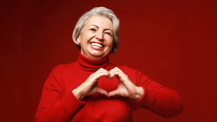 Mujer mayor con jersey rojo haciendo un corazón con las manos sobre fondo rojo liso.