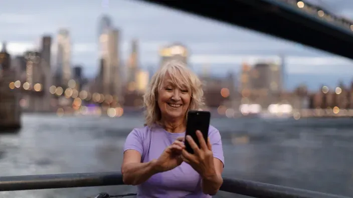 Mujer mayor utilizando un smartphone frente a un skyline urbano al atardecer.