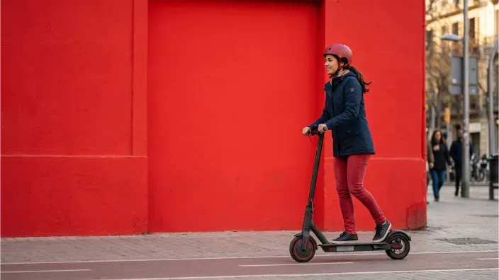 Mujer con casco circulando en patinete eléctrico por un carril bici frente a una pared roja.